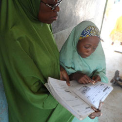 Zainab, an 11-year-old Nigerian girl, stands at the front of the class with her teacher looking at a book.