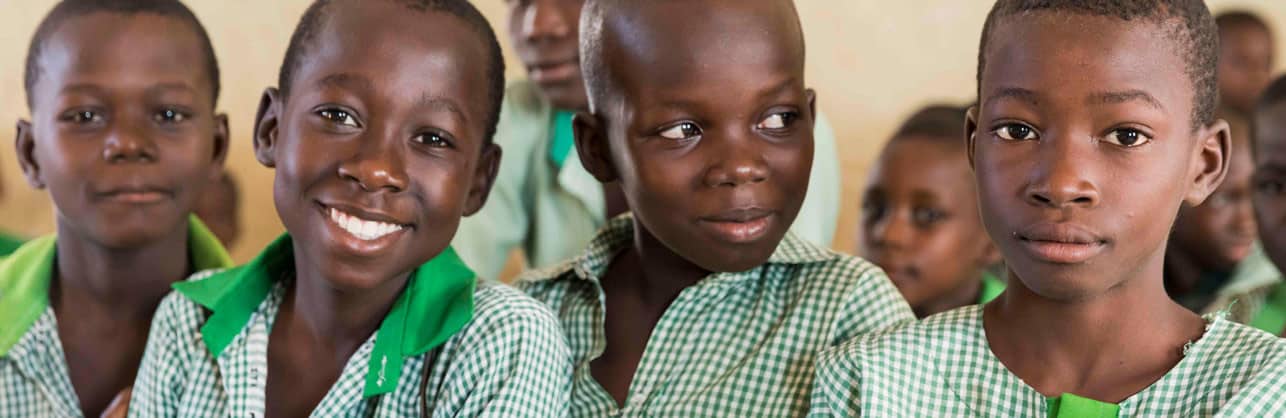 Quatro meninos nigerianos em sala de aula vestindo uniformes escolares xadrez verdes e brancos e sorrindo.