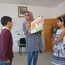 A summer reading program facilitator stands at the front of the classroom with a young Moroccan girl and boy while they read from the large book she holds.