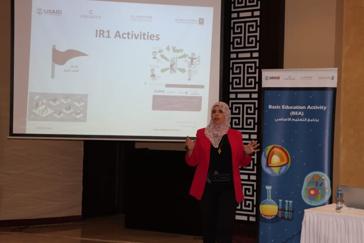 Nariman Othman, Conseiller en inclusion auprès de l'activité d'éducation de base de l'USAID (BÉA) en Cisjordanie et à Gaza, présente des activités d’inclusion devant une bannière et un écran de projection.